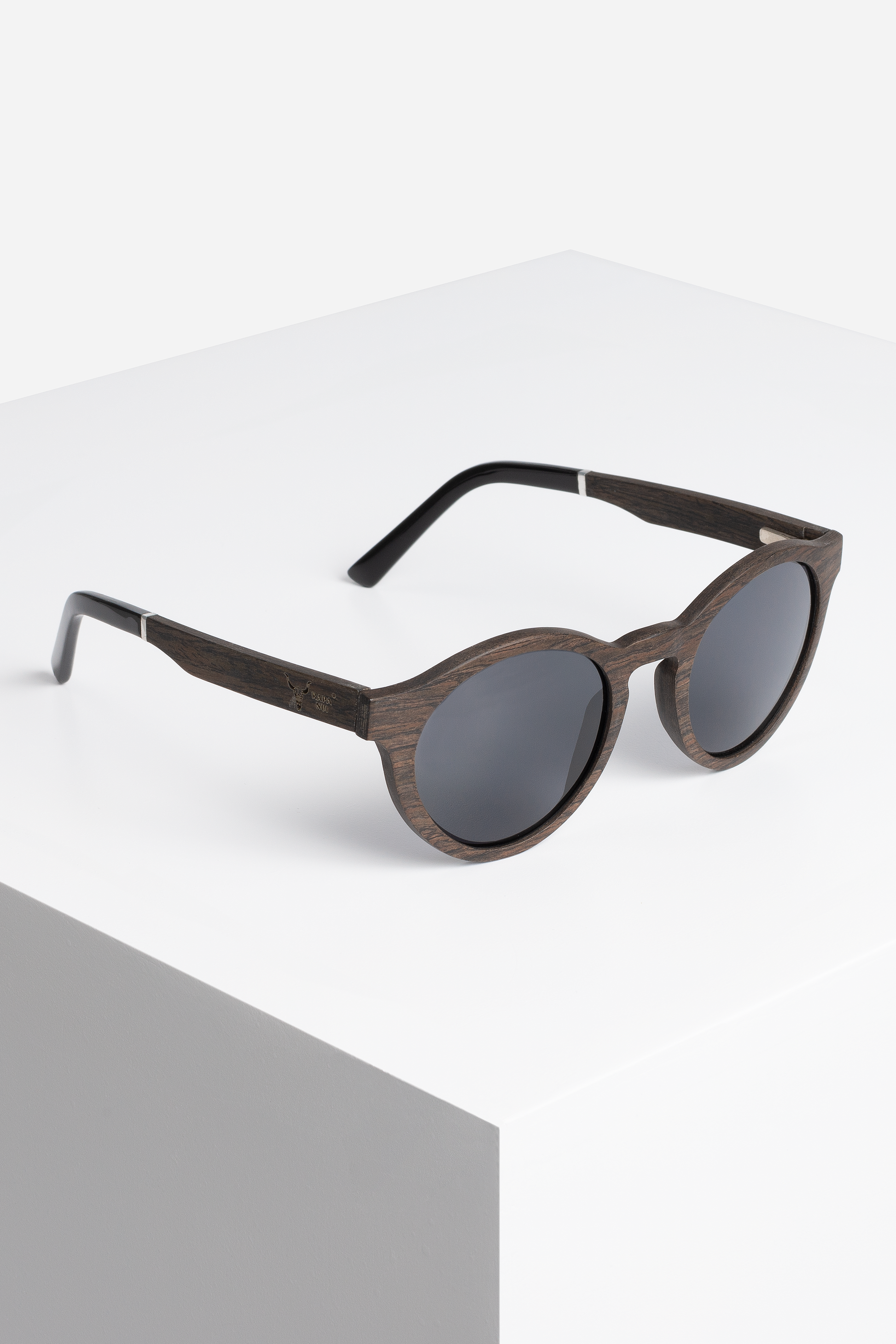 Eine stilvolle Sonnenbrille **HORIZON by RAPANUI ORIGINAL** mit runden, dunklen Gläsern und Holzrahmen mit natürlicher Holzmaserung und schwarzen Bügeln steht auf einer weißen, minimalistischen Oberfläche. Das Design strahlt eine moderne und elegante Ästhetik aus und ist gleichzeitig federleicht für angenehmen Tragekomfort.